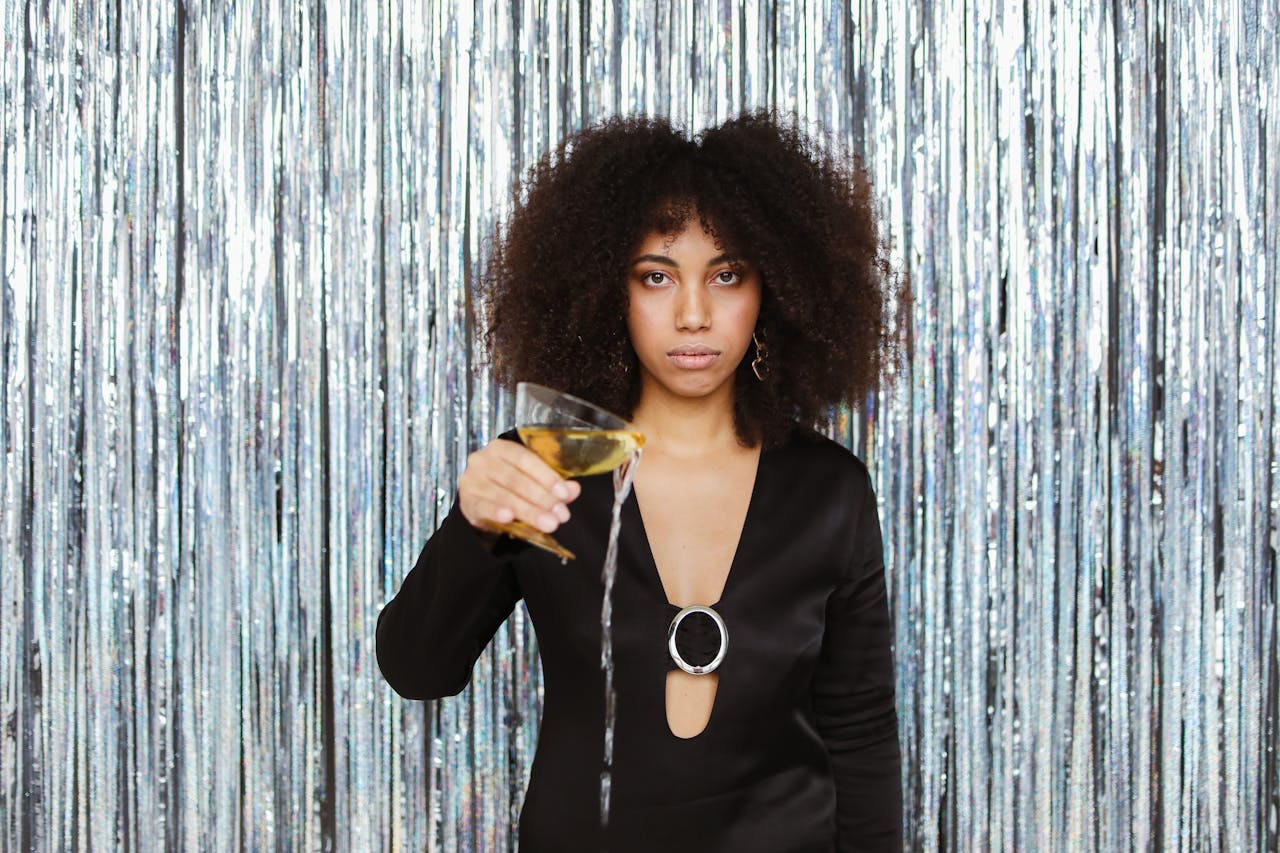 A young black woman in a black dress holds a cocktail glass, pouring it in a chic setting.